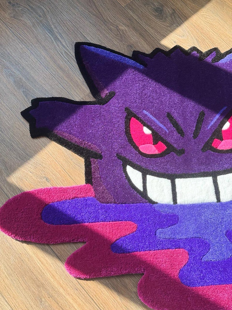 Puede incluir: Una alfombra tufting con la forma del personaje Gengar, que presenta un cuerpo de color morado intenso con un contorno negro, ojos de color rosa brillante y una amplia sonrisa blanca. La base muestra un dise&ntilde;o abstracto de charco en tonos magenta y violeta.