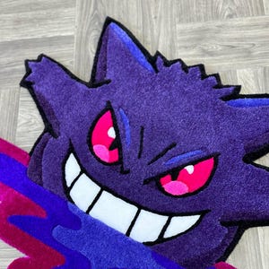 Puede incluir: Una alfombra tufting con la forma del personaje Gengar, que presenta un cuerpo de color morado intenso, orejas puntiagudas y una amplia sonrisa con dientes blancos. El personaje tiene ojos de color rosa brillante y emerge de una mancha l&iacute;quida.