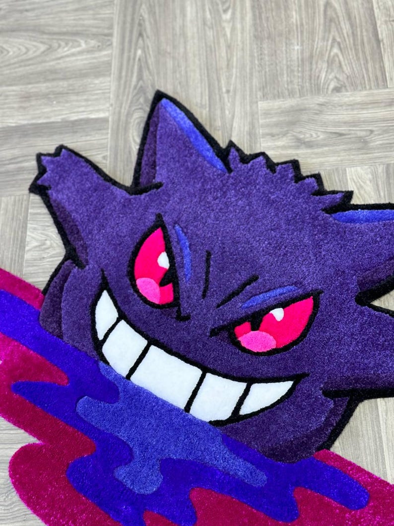 Puede incluir: Una alfombra tufting con la forma del personaje Gengar, que presenta un cuerpo de color morado intenso, orejas puntiagudas y una amplia sonrisa con dientes blancos. El personaje tiene ojos de color rosa brillante y emerge de una mancha l&iacute;quida.
