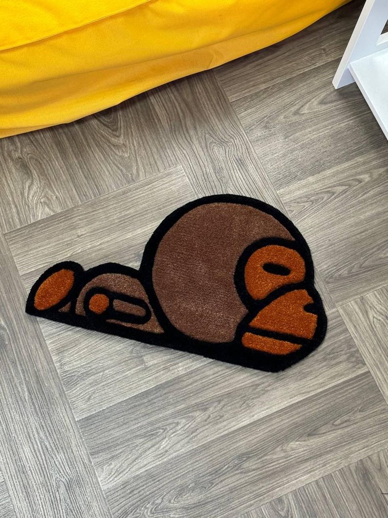 May include: A brown and black rug with a cartoon monkey design. The monkey is lying down with its head turned to the side. The rug is made of a soft, plush material.