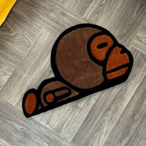 May include: A brown and orange rug with a cartoon monkey design. The monkey is lying down with its head turned to the side. The rug is on a gray wood floor.