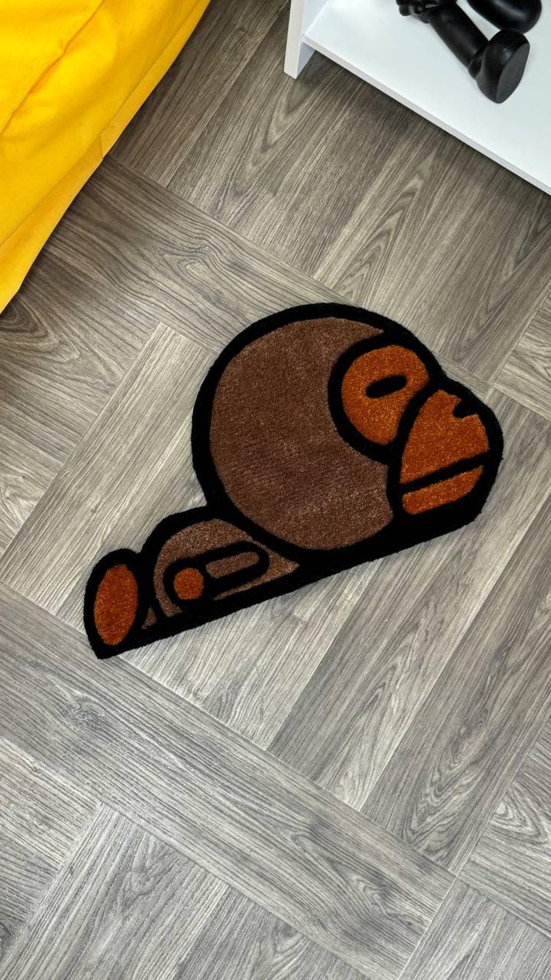 May include: A brown and orange rug with a cartoon monkey design. The monkey is lying down with its head turned to the side. The rug is on a gray wood floor.
