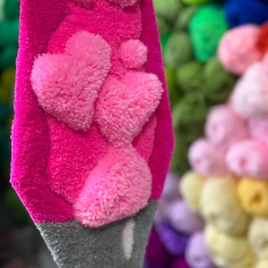 May include: A pink lava lamp made of yarn with a gray base and pink heart-shaped pom-poms. The lava lamp is hanging in front of a wall of colorful yarn pom-poms.