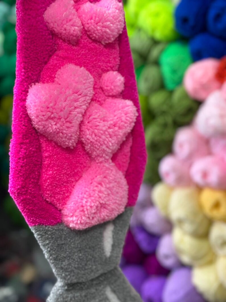May include: A pink lava lamp made of yarn with a gray base and pink heart-shaped pom-poms. The lava lamp is hanging in front of a wall of colorful yarn pom-poms.