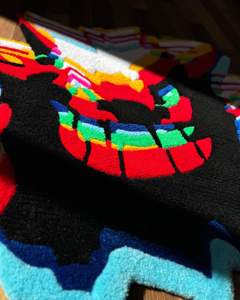 May include: A tufted rug featuring a stylized, multi-colored graphic design with a prominent black background. The pattern includes vibrant red, green, yellow, blue, and white sections with a textured, plush surface and irregular, jagged edges.