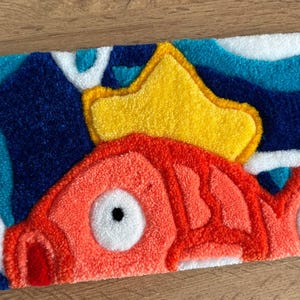 May include: A rectangular rug featuring a cartoon fish design. The fish is primarily orange with white accents and a large white eye. A yellow crown sits above the fish, set against a blue and white wave pattern background.