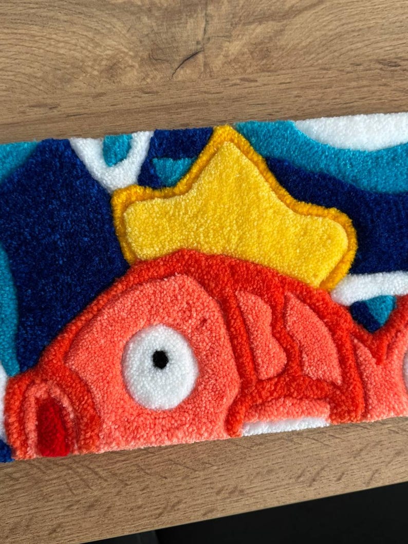 May include: A rectangular rug featuring a cartoon fish design. The fish is primarily orange with white accents and a large white eye. A yellow crown sits above the fish, set against a blue and white wave pattern background.