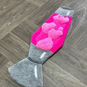 May include: A pink and gray rug shaped like a lava lamp. The lamp has a pink, fuzzy texture and a gray base.