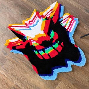 May include: A tufted rug featuring a stylized, abstract character design with a large black central area, vibrant red, orange, yellow, and white jagged accents at the top, and a bright blue, green, and multicolored layered border at the bottom.