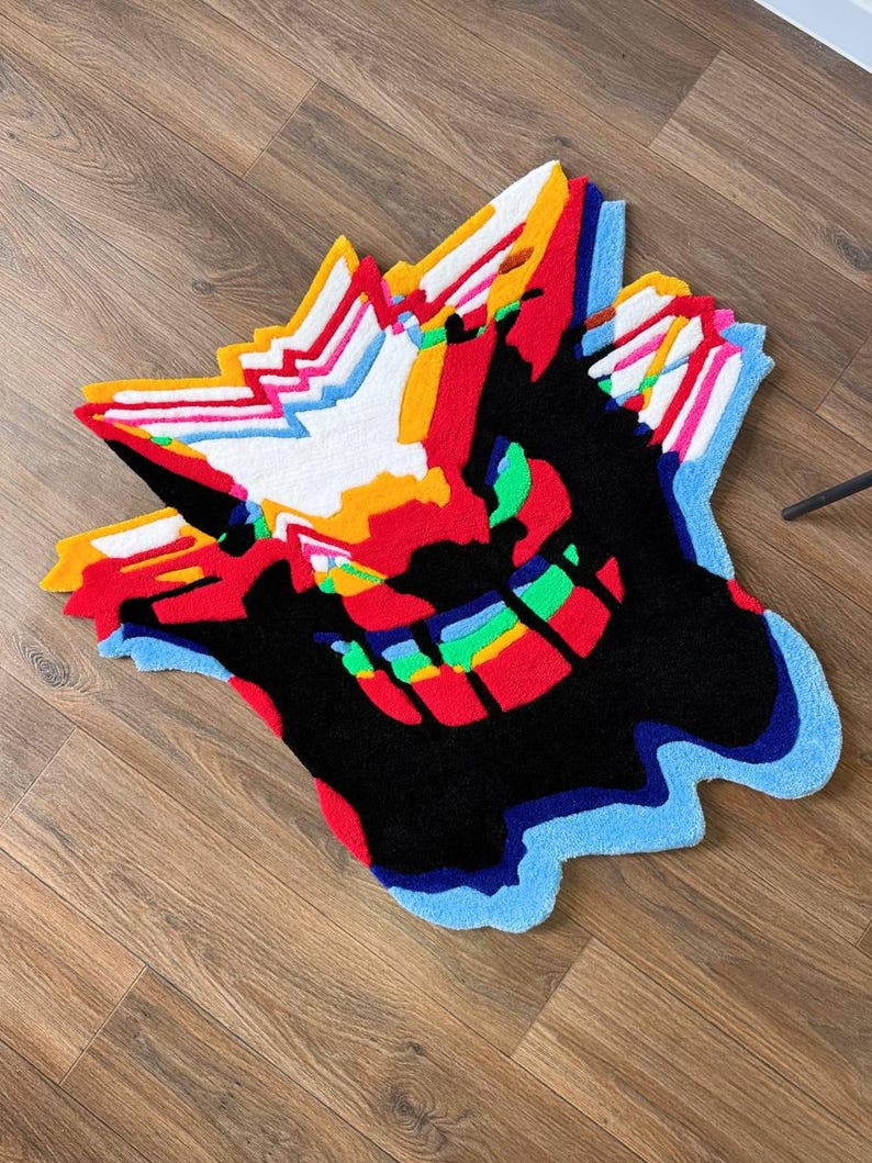 May include: A tufted rug featuring a stylized, abstract character design with a large black central area, vibrant red, orange, yellow, and white jagged accents at the top, and a bright blue, green, and multicolored layered border at the bottom.