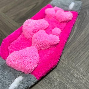 May include: A pink and gray rug with a heart design. The rug is made of fluffy yarn and has a textured surface.