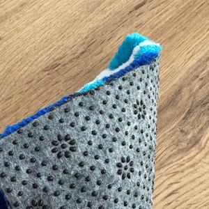 May include: Close-up of a section of a colorful rug with a blue, white, and yellow design. The underside of the rug is gray with black, flower-shaped anti-slip grips. The rug is on a wooden surface.