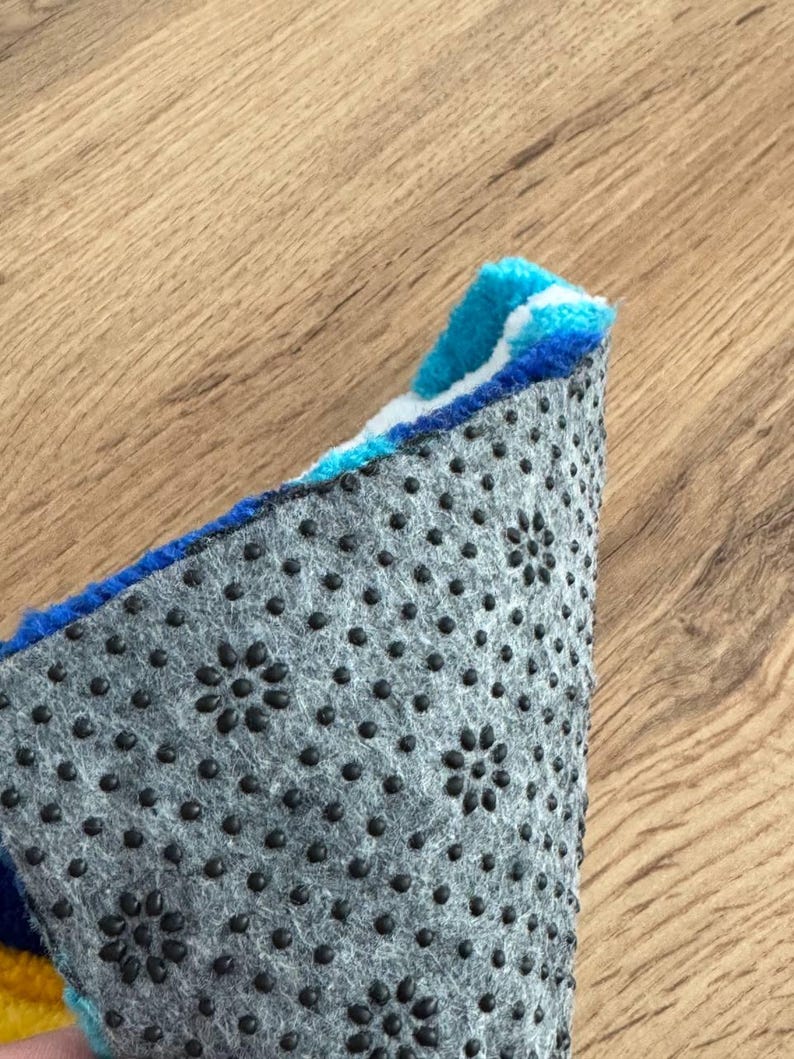 May include: Close-up of a section of a colorful rug with a blue, white, and yellow design. The underside of the rug is gray with black, flower-shaped anti-slip grips. The rug is on a wooden surface.