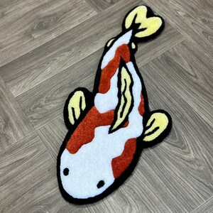 May include: A white and orange koi fish rug with yellow fins. The rug is made of fluffy, textured material and has a black outline.