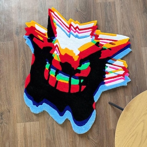 May include: A tufted rug featuring a stylized, glitch-effect graphic of a creature face. The design incorporates vibrant layers of red, yellow, white, blue, green, and pink, all set against a dominant black background with a melting, irregular edge.