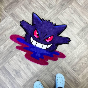 Puede incluir: Una alfombra de mechones con la forma del personaje morado Gengar, con orejas puntiagudas, ojos rojos brillantes y una amplia sonrisa blanca. El personaje parece emerger de un charco l&iacute;quido de color azul oscuro y magenta.