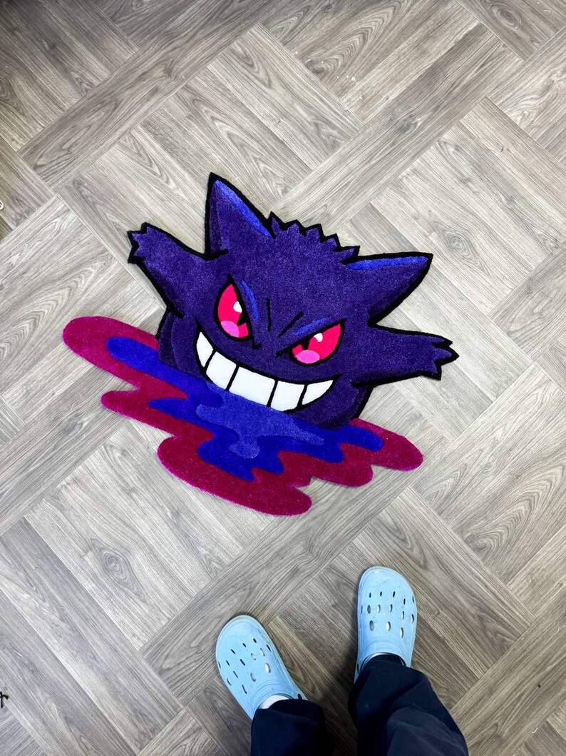 Puede incluir: Una alfombra de mechones con la forma del personaje morado Gengar, con orejas puntiagudas, ojos rojos brillantes y una amplia sonrisa blanca. El personaje parece emerger de un charco l&iacute;quido de color azul oscuro y magenta.