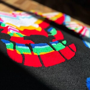 May include: A black tufted rug featuring a vibrant, multi-colored graphic design. The surface displays bold, raised patterns in red, yellow, green, blue, and white, creating a textured, high-contrast appearance with distinct, thick yarn loops.