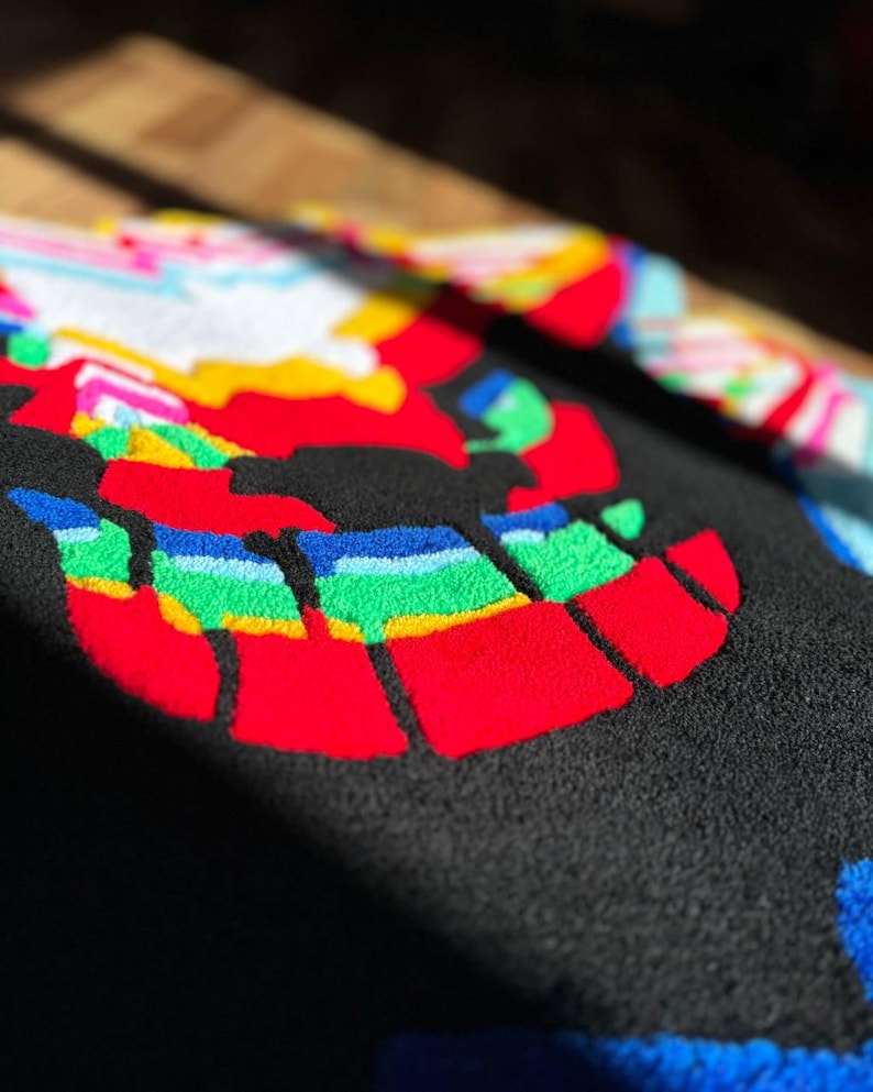 May include: A black tufted rug featuring a vibrant, multi-colored graphic design. The surface displays bold, raised patterns in red, yellow, green, blue, and white, creating a textured, high-contrast appearance with distinct, thick yarn loops.