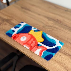 May include: A rectangular, colorful rug featuring a cartoon fish, a yellow star, and abstract blue and white shapes. The fish is primarily coral with white accents. The rug is placed on a wooden surface.