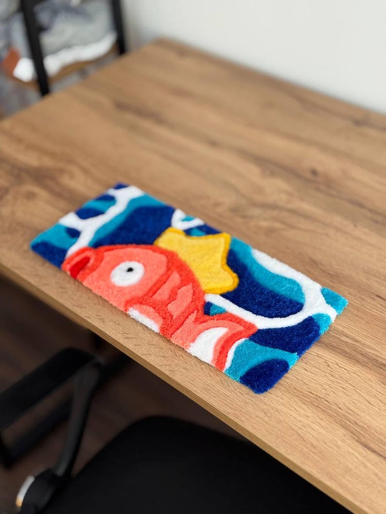 May include: A rectangular, colorful rug featuring a cartoon fish, a yellow star, and abstract blue and white shapes. The fish is primarily coral with white accents. The rug is placed on a wooden surface.