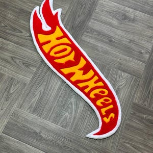 May include: A red and yellow Hot Wheels logo rug with a white outline. The rug is shaped like a flame and has a fuzzy texture.