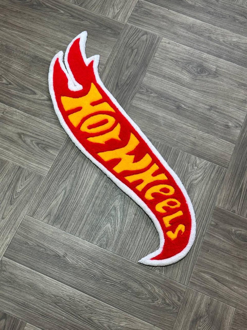 May include: A red and yellow Hot Wheels logo rug with a white outline. The rug is shaped like a flame and has a fuzzy texture.