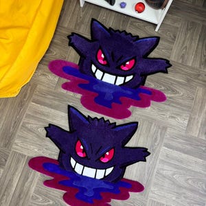 Puede incluir: Dos alfombras de tufting con la forma de la criatura fantasma morada Gengar, con ojos rojos brillantes, una amplia sonrisa blanca y una base de charco l&iacute;quido en morado oscuro y magenta, sobre un suelo de madera con una estanter&iacute;a blanca al fondo.