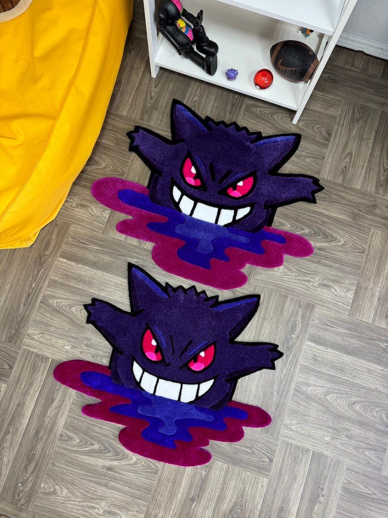 Puede incluir: Dos alfombras de tufting con la forma de la criatura fantasma morada Gengar, con ojos rojos brillantes, una amplia sonrisa blanca y una base de charco l&iacute;quido en morado oscuro y magenta, sobre un suelo de madera con una estanter&iacute;a blanca al fondo.