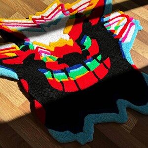 May include: A tufted rug featuring a stylized creature design with a large black silhouette, a wide grin with rainbow-colored teeth, and vibrant red, yellow, white, and light blue accents. The rug has an irregular, organic shape with textured fibers.