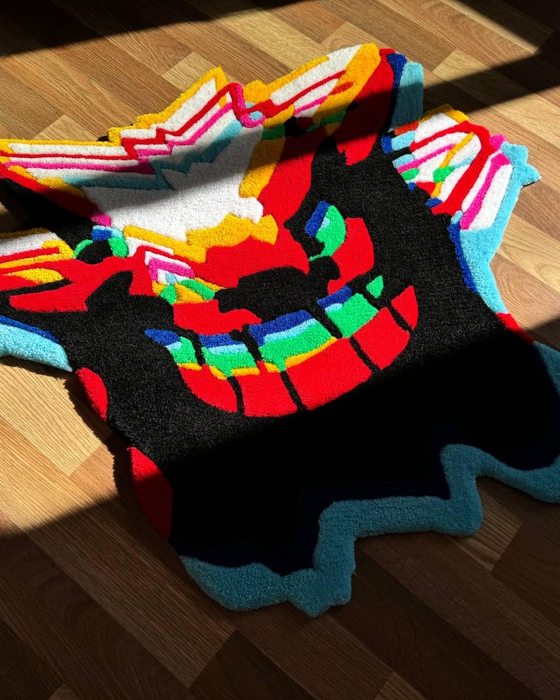 May include: A tufted rug featuring a stylized creature design with a large black silhouette, a wide grin with rainbow-colored teeth, and vibrant red, yellow, white, and light blue accents. The rug has an irregular, organic shape with textured fibers.