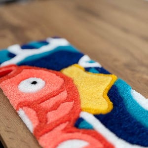 May include: A colorful, rectangular rug featuring a stylized fish design. The rug has a coral-colored fish with a black eye, a yellow shape, and blue and white wave patterns. The rug is made of a soft, textured material.