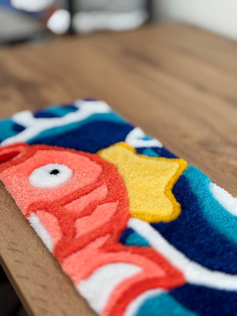 May include: A colorful, rectangular rug featuring a stylized fish design. The rug has a coral-colored fish with a black eye, a yellow shape, and blue and white wave patterns. The rug is made of a soft, textured material.