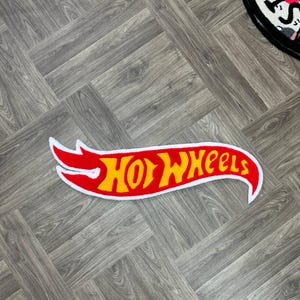 May include: A red and yellow Hot Wheels logo rug with a white outline. The rug is shaped like a racing flag and has a white border.