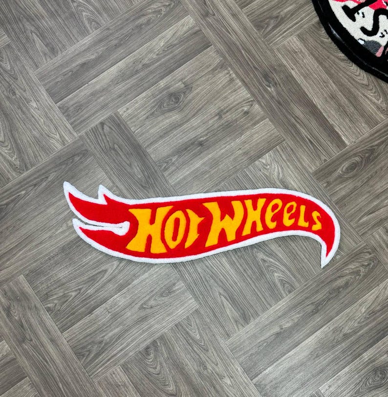 May include: A red and yellow Hot Wheels logo rug with a white outline. The rug is shaped like a racing flag and has a white border.