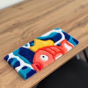May include: A rectangular, colorful rug featuring a cartoon fish design. The rug has a blue, white, and yellow background with a large red fish. The rug is made of a soft, textured material.