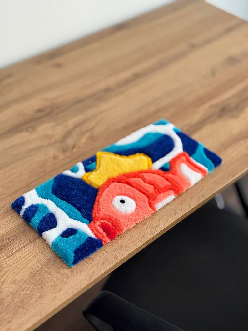 May include: A rectangular, colorful rug featuring a cartoon fish design. The rug has a blue, white, and yellow background with a large red fish. The rug is made of a soft, textured material.