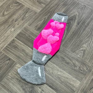 May include: A pink and gray lava lamp rug with a fuzzy texture. The lamp is shaped like a traditional lava lamp with a round base and a tall, thin neck.