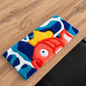 May include: A rectangular rug featuring a cartoon fish design. The rug has a red fish with a yellow crown, set against a blue and white wavy background. The rug is placed on a wooden surface.