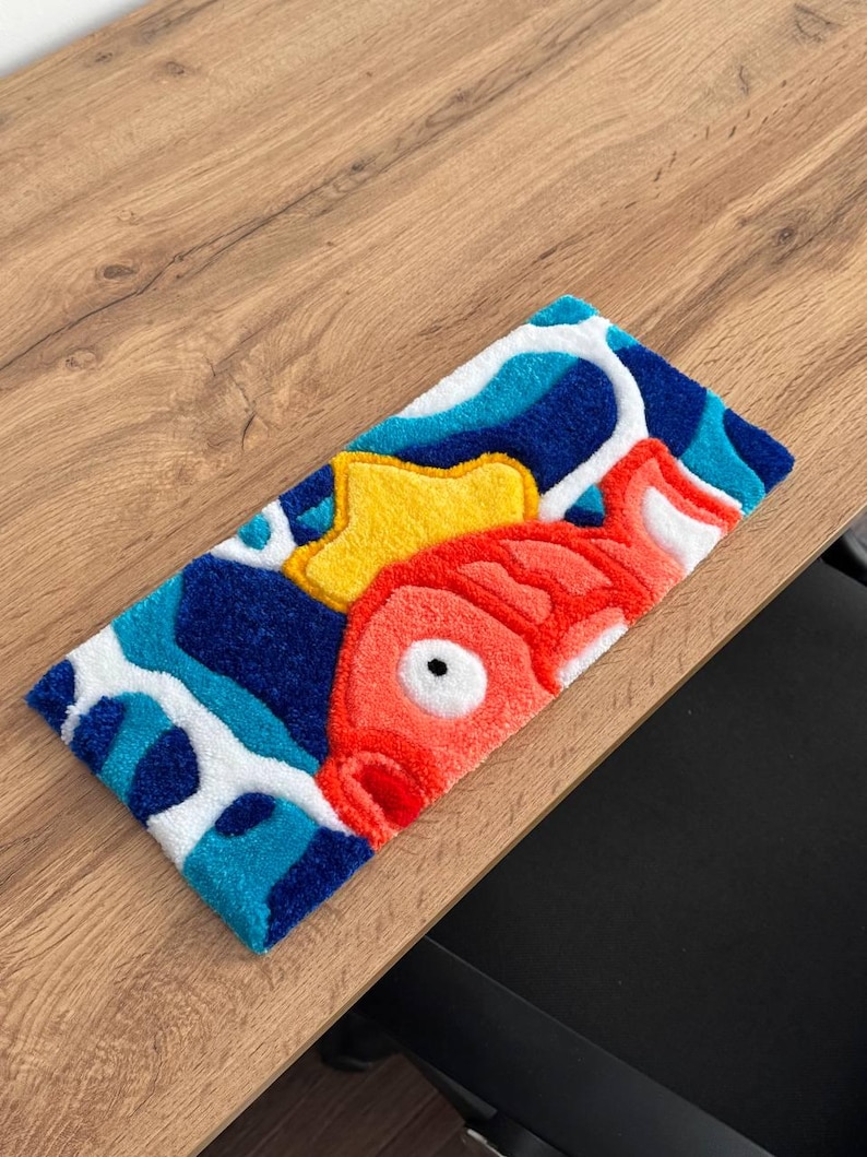 May include: A rectangular rug featuring a cartoon fish design. The rug has a red fish with a yellow crown, set against a blue and white wavy background. The rug is placed on a wooden surface.