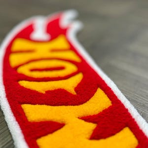 May include: A red and yellow Hot Wheels logo patch with a white border. The logo is made of a fuzzy material and is shaped like a flame.