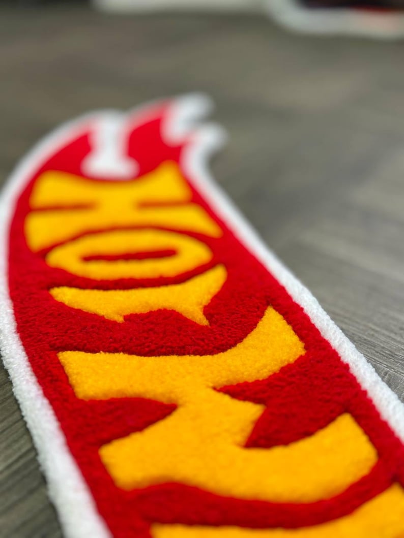 May include: A red and yellow Hot Wheels logo patch with a white border. The logo is made of a fuzzy material and is shaped like a flame.