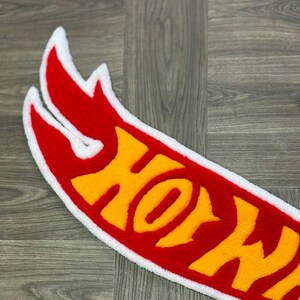 May include: A red and yellow Hot Wheels logo rug with a white border. The logo is made of fluffy, plush material.