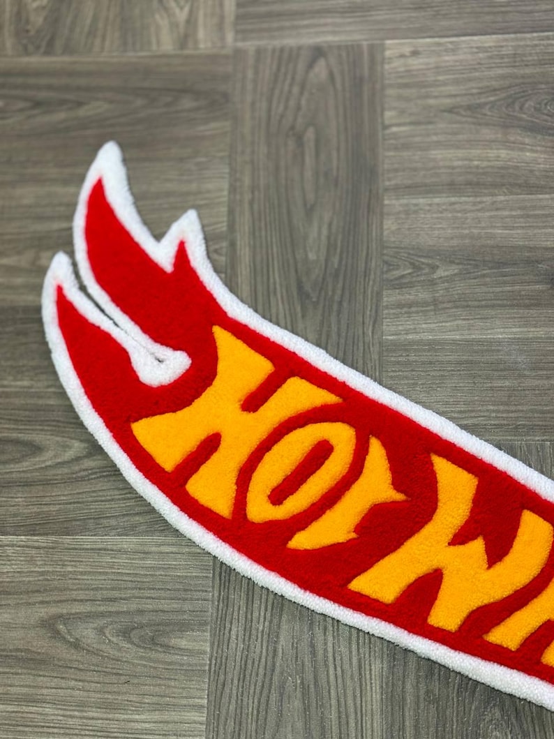 May include: A red and yellow Hot Wheels logo rug with a white border. The logo is made of fluffy, plush material.