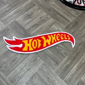 May include: A red and yellow "Hot Wheels" logo rug with a white border. The rug is shaped like a flame and is on a wood floor.