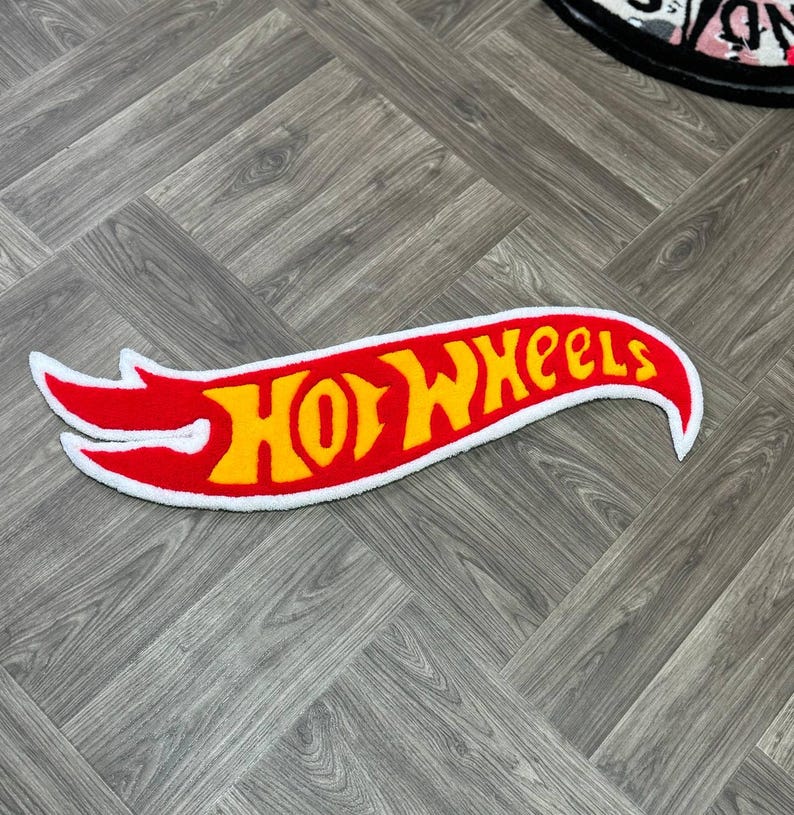 May include: A red and yellow "Hot Wheels" logo rug with a white border. The rug is shaped like a flame and is on a wood floor.