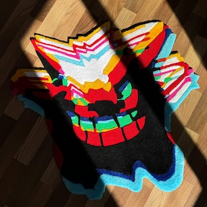 May include: A tufted rug featuring a stylized, multi-layered graphic design with vibrant colors including red, black, white, yellow, light blue, and green. The rug has an irregular, jagged shape with overlapping patterns that create a visual depth effect.