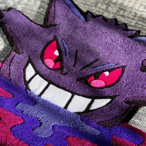 Puede incluir: Una alfombra de mechones que presenta al personaje morado Gengar con orejas puntiagudas, ojos rojos afilados y una amplia sonrisa blanca. El personaje est&aacute; rodeado por una base l&iacute;quida arremolinada en morado y magenta con acabado de felpa.