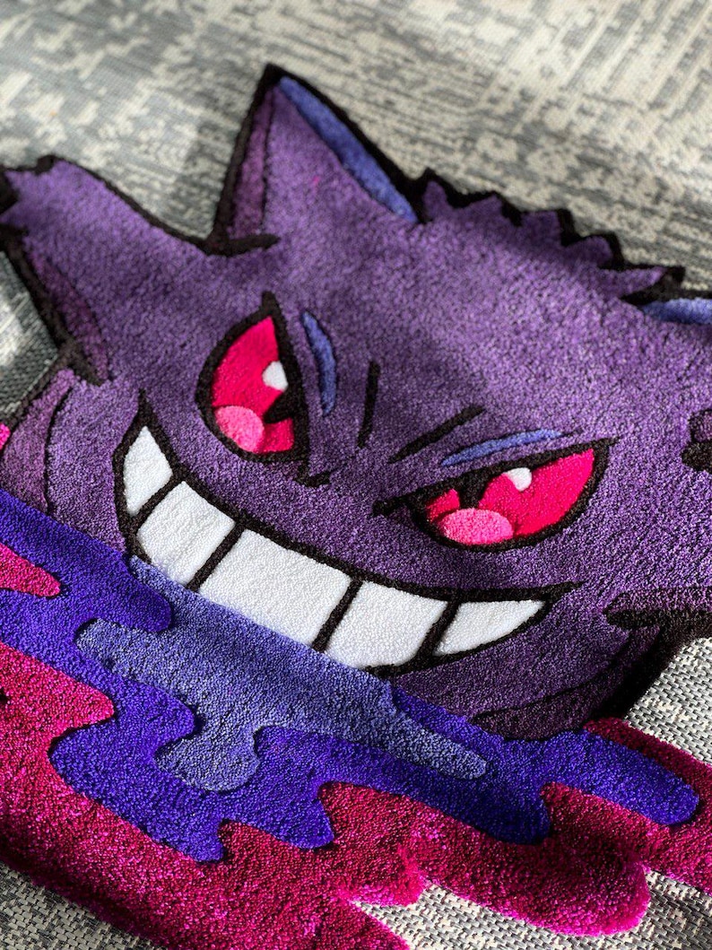 Puede incluir: Una alfombra de mechones que presenta al personaje morado Gengar con orejas puntiagudas, ojos rojos afilados y una amplia sonrisa blanca. El personaje est&aacute; rodeado por una base l&iacute;quida arremolinada en morado y magenta con acabado de felpa.
