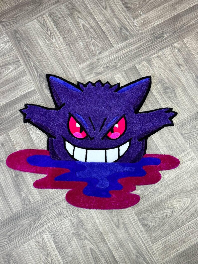 Puede incluir: Una alfombra tufting con la forma del personaje Gengar, que presenta un cuerpo de color morado intenso, orejas puntiagudas y una amplia sonrisa con dientes blancos. El personaje tiene ojos rojos brillantes y emerge de un charco de formas l&iacute;quidas azules y magenta.